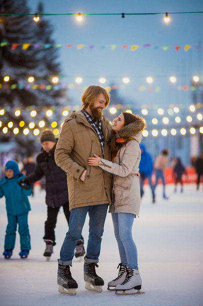 Young couple in love Caucasian man with blond hair with long hair and beard and beautiful woman have fun, active date ice skating on the ice arena in the evening city square in winter on Christmas Eve - 写真・画像