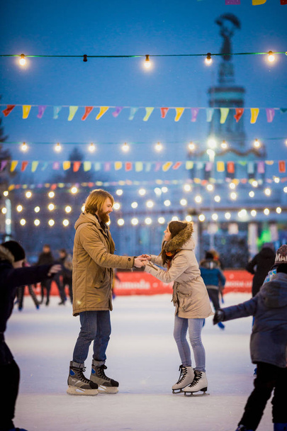 若いカップルが長い髪の金髪の白人男性を愛し、ひげと美しい女性がクリスマスイブに冬の夜市広場でアイス アリーナでスケートを楽しみ、アクティブな日付氷を持って - 写真・画像