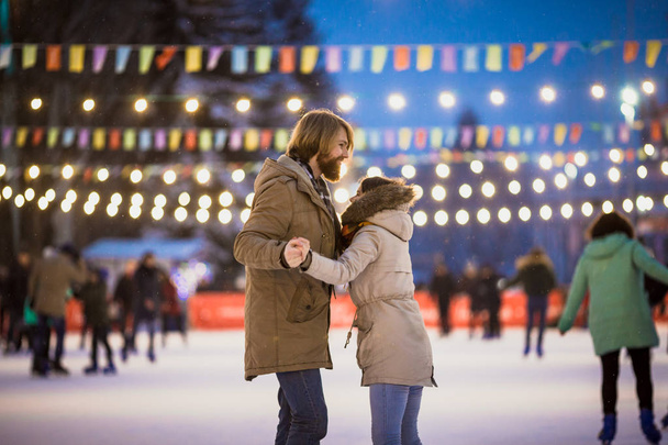 Jovem casal apaixonado caucasiano com cabelo loiro com cabelos longos e barba e mulher bonita se divertir, data ativa patinação no gelo na arena de gelo na praça da cidade à noite no inverno na véspera de Natal
 - Foto, Imagem