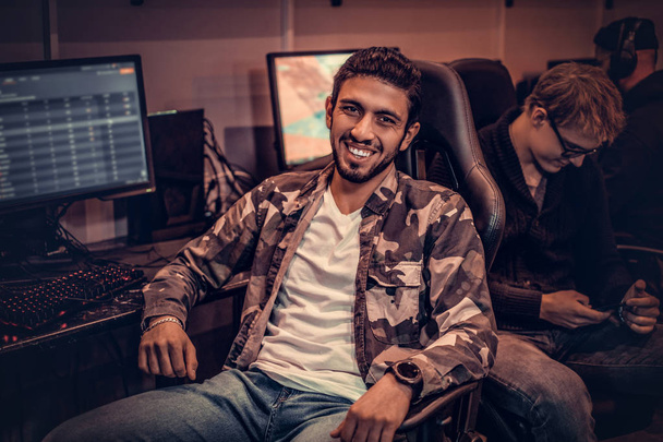 Nuori iloinen intialainen mies yllään armeijan paita istuu gamer tuoli ja katsot kameraa pelaamista klubi tai internet kahvila
. - Valokuva, kuva