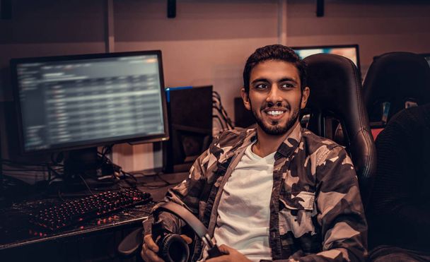Een jonge vrolijke Indiase kerel het dragen van een militaire shirt zittend op een gamer stoel in een gaming club of internet café. - Foto, afbeelding