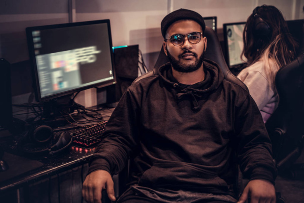 African American bebaarde man dragen hoodie en dop op een gamer stoel zitten en kijken naar een camera in een gaming club of internet café. - Foto, afbeelding