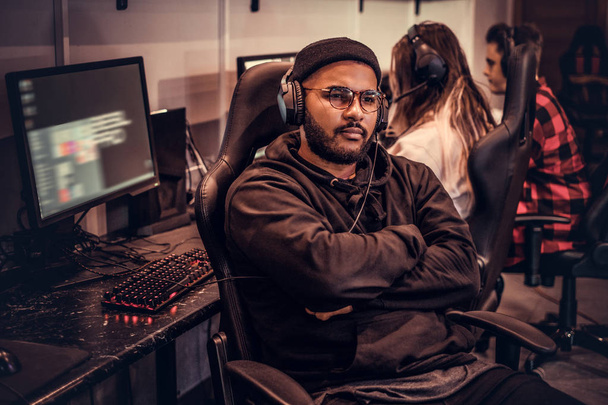 Afro-Amerikaanse bebaarde man dragen hoodie en pet met haar armen gekruist zittend op een gamer stoel in een gaming club of internet café. - Foto, afbeelding