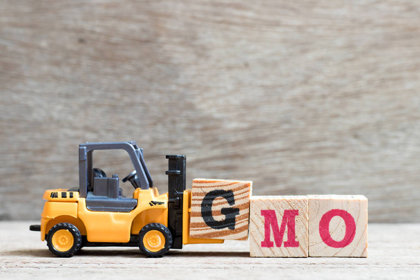 Spielzeuggabelstapler halten Buchstabenblock g, um das Wort gmo (Abkürzung für genetisch veränderte Organismen) auf Holzgrund zu vervollständigen - Foto, Bild