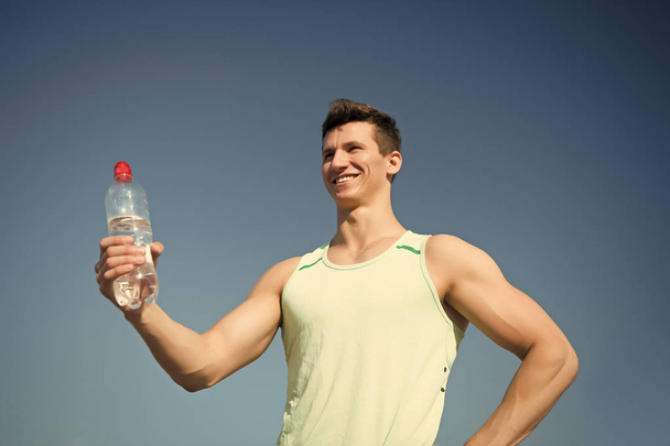 Athlete smiling in green tshirt on blue sky. Man with water bottle in muscular hands. Sport and fitness concept. Bodybuilder with muscles biceps, triceps on sunny day. Healthy lifestyle and health. - Fotoğraf, Görsel