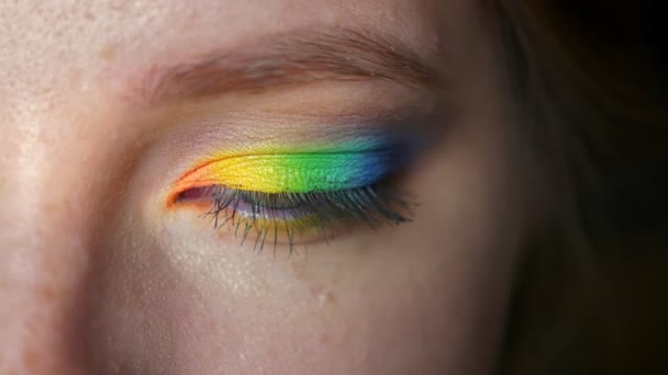 Rainbow makeup on closeup blue eye of caucasian girl looking at camera and showing colorful eyeshadows, black background - Footage, Video