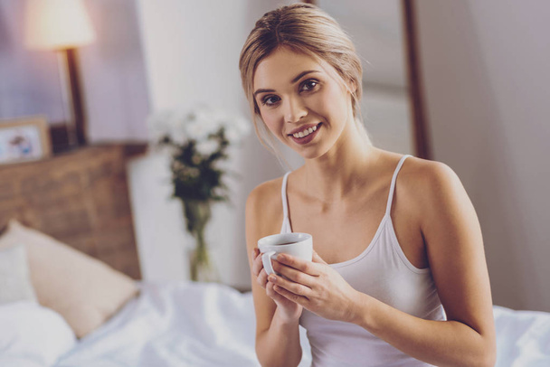Coffee lover. Beautiful young woman posing with a cup of coffee and smiling pleasantly while sitting on the bed - Φωτογραφία, εικόνα