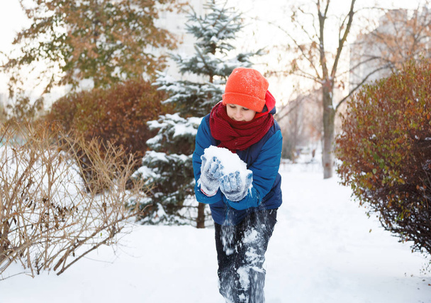cute young boy in red hat blue jacket holds and plays with snow, has fun, smiles, makes snowman in winter park. Active lifestyle, winter activity, outdoor winter games, snowballs.  - Photo, Image