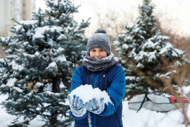 lindo joven con sombrero rojo chaqueta azul sostiene y juega con la nieve, se divierte, sonríe, hace muñeco de nieve en el parque de invierno. Estilo de vida activo, actividad invernal, juegos al aire libre de invierno, bolas de nieve
.  - Foto, imagen