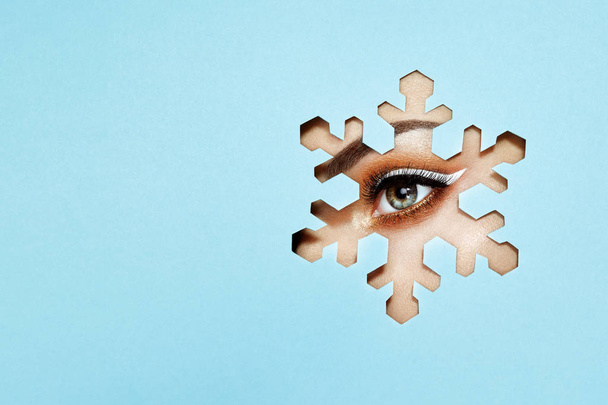 The Eyes of the Young Beautiful Woman with Bright Golden Shadows and Expressive Eyebrows, Looks in the Snowflake Pattern out of Colored Paper. Snowflake. Christmas Patterns. Blue Paper - Фото, изображение
