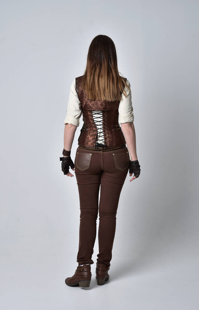 retrato de comprimento total da menina morena vestindo roupas de couro marrom steampunk. postura de pé com as costas para a câmera no fundo do estúdio cinza
. - Foto, Imagem
