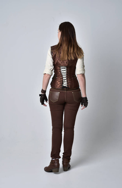 Pełna długość Portret brunetka dziewczyna ubrana w strój steampunk brązowej skóry. Stojący poza z powrotem do aparatu na studio szary tło. - Zdjęcie, obraz