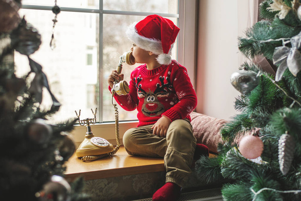 Мальчик в кепке Санта-Клауса сидит на окне и звонит Санте
. - Фото, изображение