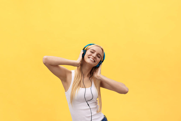 Lifestyle Concept: Portrait of a joyful woman in white t-shirt and listening to music with earphones while dancing and singing isolated over yellow background - Fotó, kép