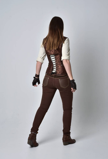 Ολόσωμο πορτρέτο μελαχρινή κοπέλα φορώντας στολή steampunk καφέ δέρμα. θέτουν στέκεται με την πλάτη στην κάμερα σε φόντο γκρι στούντιο. - Φωτογραφία, εικόνα