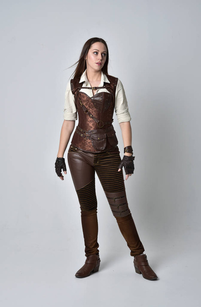 retrato de longitud completa de chica morena con traje steampunk de cuero marrón. pose de pie sobre fondo gris estudio
. - Foto, imagen