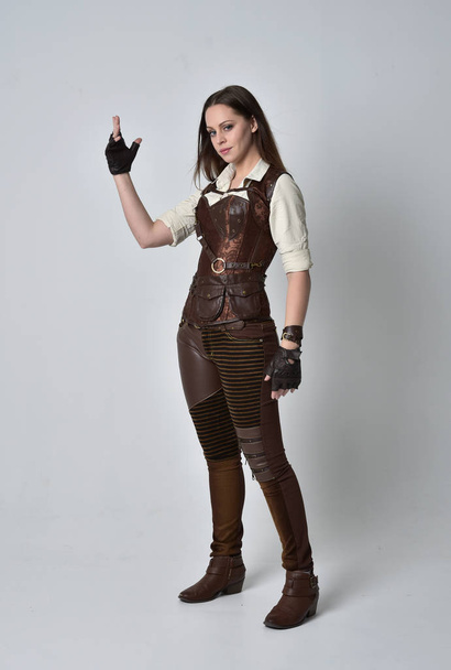po celé délce portrét bruneta dívka, která nosí hnědé kožené steampunk oblečení. představovat stojící na šedé studio pozadí. - Fotografie, Obrázek