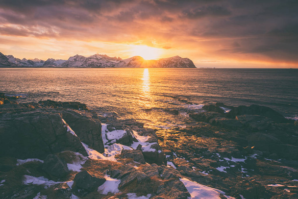Amazing colors of north atlantic sunset, winter seascape with ocean, rocky seacoast, beautiful cloudy sky, snowy mountains and sun on background, Vikten, Lofoten Islands, Northern Norway - Photo, Image