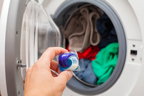 Close up picture of person holding laundry detergent 3 in 1 pod capsule, washing machine with laundry in the background - Photo, image