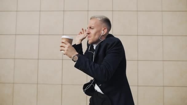 Happy young businessman with coffee listening to music in headphones on smartphone and funny dancing near office building. Funny dancing moves. Slow motion - Materiał filmowy, wideo
