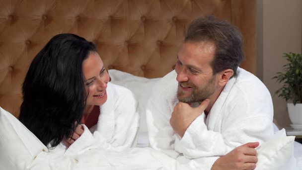 Mature married couple in bathrobes talking, lying in bed at the hotel together - Photo, Image