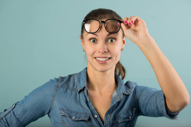 beautiful young girl in a denim shirt with glasses on a blue background with astonishment on her face - Foto, Imagen