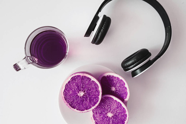 Верхний вид композиции напитка в прозрачной стеклянной чашке, нарезанные грейпфруты и белые минималистские наушники на белой поверхности с измененными цветами, чтобы создать абстрактный ультрафиолетовый вид
 - Фото, изображение