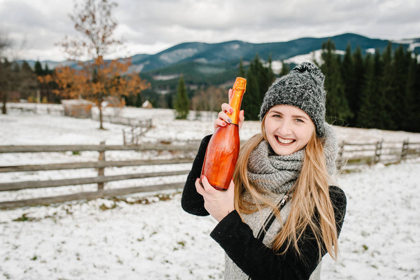 Cheerful woman holding bottle of champagne on winter mountains background - 写真・画像