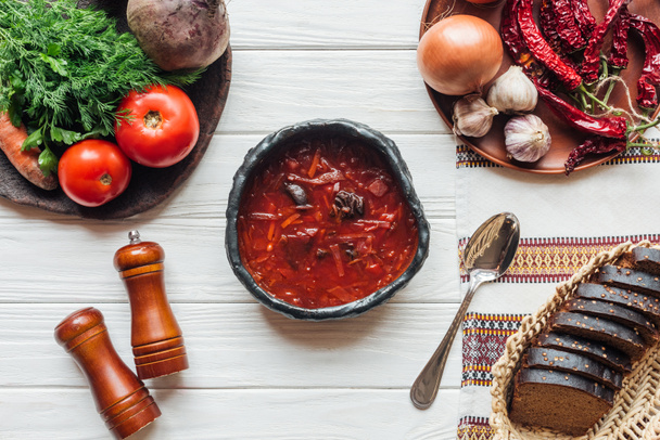 Draufsicht auf köstliche traditionelle Rote-Bete-Suppe mit Besteck, Zutaten und Roggenbrot auf weißem Holzgrund - Foto, Bild