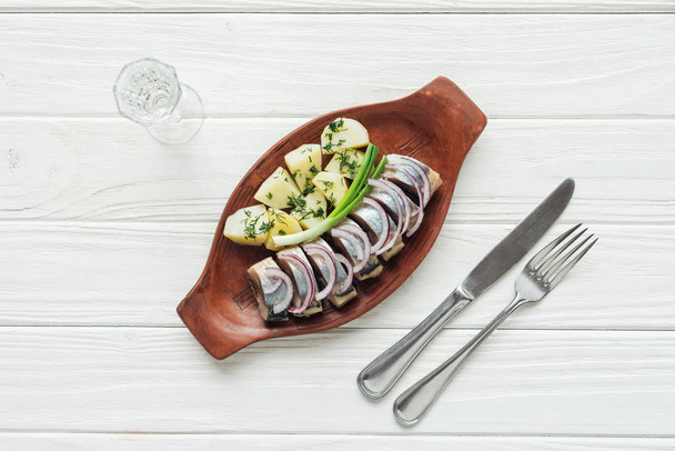 top view of marinated herring, potatoes and onions in earthenware plate with glass of vodka and cutlery on white wooden background - Photo, Image
