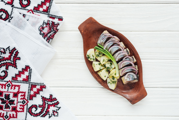 top view of marinated herring with potatoes and onions in earthenware plate with embroidered towel on white wooden background - Photo, Image