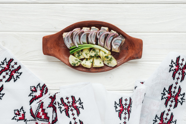 heerlijk gemarineerde haring met aardappelen en uien in aardewerk plaat met geborduurde handdoek op witte houten achtergrond - Foto, afbeelding