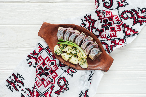 traditioneel gemarineerde haring met aardappelen en uien in aardewerk plaat met geborduurde handdoek op witte houten achtergrond - Foto, afbeelding
