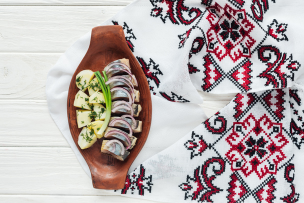 delicious marinated herring with potatoes and onions in earthenware plate with embroidered towel on white wooden background - Fotó, kép
