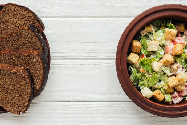 вид зверху смачний традиційний салат Цезаря з грінками в мисці і житній хліб на білому дерев'яному фоні
 - Фото, зображення