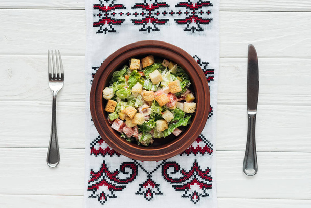 Beyaz ahşap zemin üzerinde lezzetli geleneksel Sezar salatası kruton, çatal bıçak takımı ve işlemeli havlu üstten görünüm - Fotoğraf, Görsel