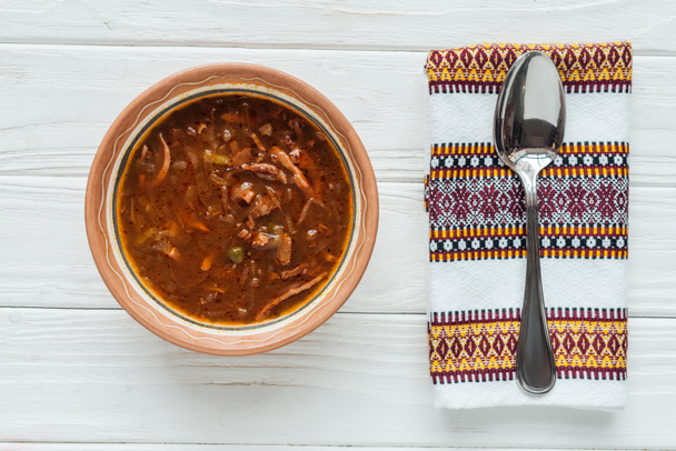 Beyaz ahşap zemin üzerine kaşık ve işlemeli havlu ile lezzetli geleneksel karışık et çorbası üstten görünüm - Fotoğraf, Görsel