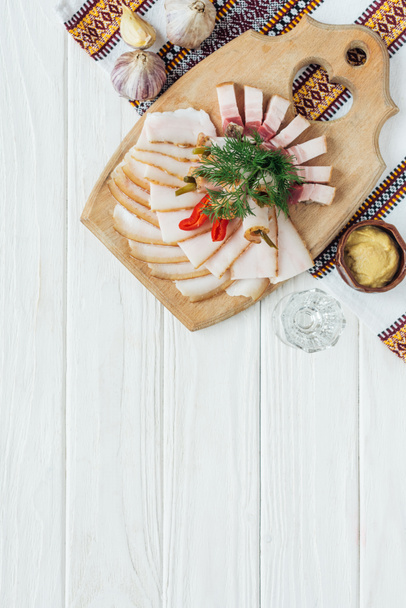 traditional sliced smoked lard on cutting board with mustard, embroidered towel and glass of vodka on white wooden background - Фото, зображення