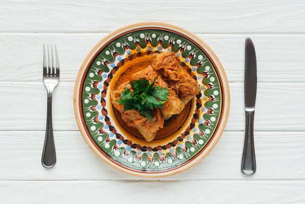 κάτοψη του νόστιμο γεμιστό λάχανο ρολά με μαϊντανό στο πιάτο, μαχαιροπίρουνα και λευκό φόντο ξύλινη - Φωτογραφία, εικόνα