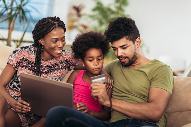 Ładna dziewczynka afro-amerykański i jej pięknych młodych rodziców za pomocą laptopa i robi zakupy online, siedząc na kanapie w domu. - Zdjęcie, obraz