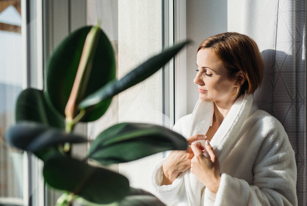 A young woman with bath robe and closed eyes standing indoors by a window in the morning, meditating. Copy space. - Foto, Imagen