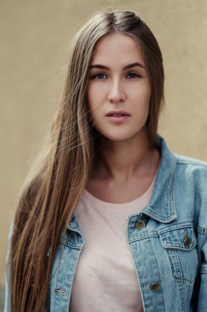 Young beautiful girl with long hair in a T-shirt and denim jacket on the background of vintage concrete wall. - Фото, зображення
