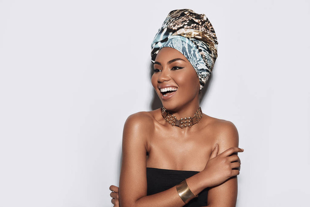Picture of a goddess. Attractive young African woman in turban smiling while standing against grey background - Fotó, kép