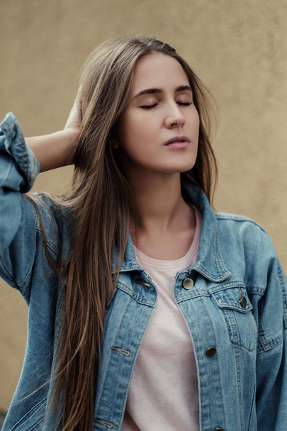 Young beautiful girl with long hair in a T-shirt and denim jacket on the background of vintage concrete wall. - Foto, afbeelding