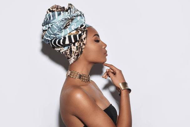 Prideful woman. Side view of attractive young African woman in turban keeping hand on chin while standing against grey background - Fotoğraf, Görsel