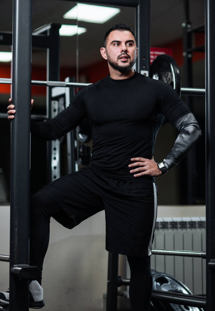 Handsome with big muscles in black clothes posing next to the barbell - Foto, afbeelding