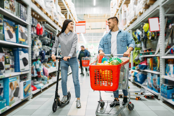 Coppia in negozio, marito con carrello pieno di merci e moglie sullo scooter. Uomo e donna in un supermercato, shopping di famiglia
 - Foto, immagini