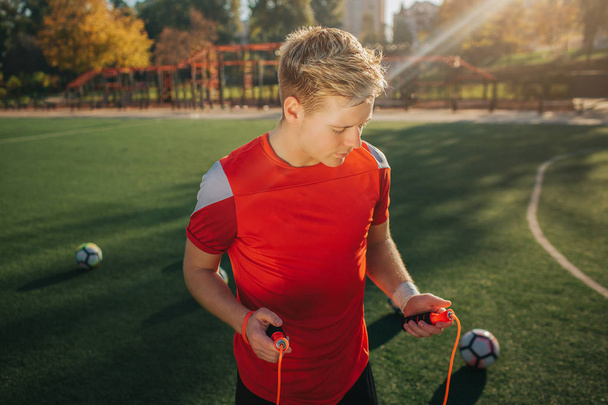 Blonde young man holding jump rope iin hands. He looks at it. Guy stand on lawn outside. Sun is shining. Balls are all around young man. - Foto, afbeelding