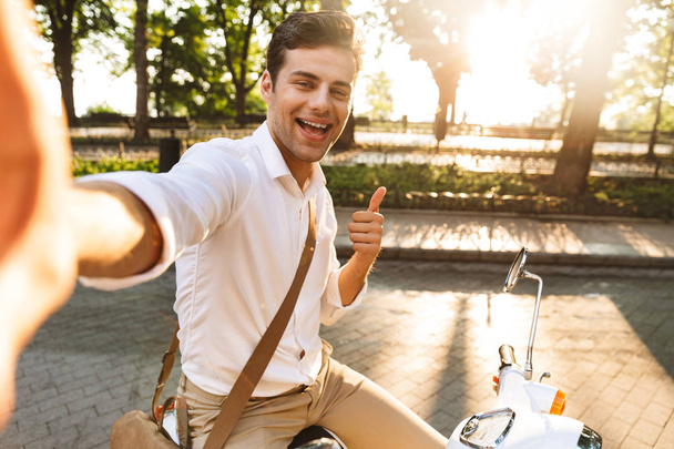 Photo of a handsome excited young business man walking outdoors on scooter take a selfie by camera. - Photo, Image