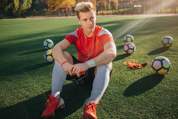 Serious and concentrated young player sit on lawn and look to right. He listen to music through headphones. Guy hold phone in hands. Balls are behind him. - Foto, afbeelding